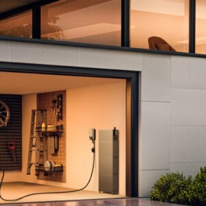 מכונית חשמלית שחורה נטענת במוסך מודרני המופעל על ידי ערכת סולאר היברידית חד-פאזית בהספק 6 קילוואט עם מערכת אחסון EcoFlow Power Ocean בהספק 5 קילוואט-שעה ולוחות פוטו-וולטאיים בהספק 3300 וואט, המציגה תשתית מתקדמת לאנרגיה מתחדשת; כלים מסודרים בקפידה מקשטים את הקיר, בעוד חשמל המופק מאנרגיה סולארית מאיר את חדר השינה הסמוך הנראה מבעד לחלון מעל.